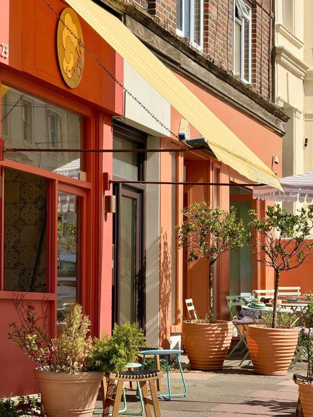 Un bâtiment à la façade rouge, avec un auvent jaune et des places assises à l’extérieur, agrémentées de plantes en pot.