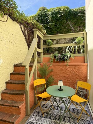 Zona exterior petita amb taula i cadires, una escala i una paret amb plantes.