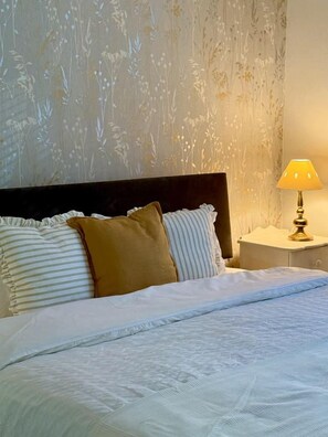 Ein Bett mit weißer Bettwäsche, einem braunen Kissen und gestreiften Kissen. Rechts daneben ein Nachttisch mit einer Lampe.