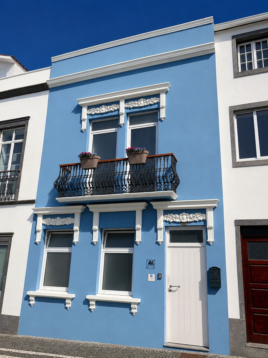 Un edificio de dos pisos con una puerta blanca, un balcón con macetas y una fachada azul.