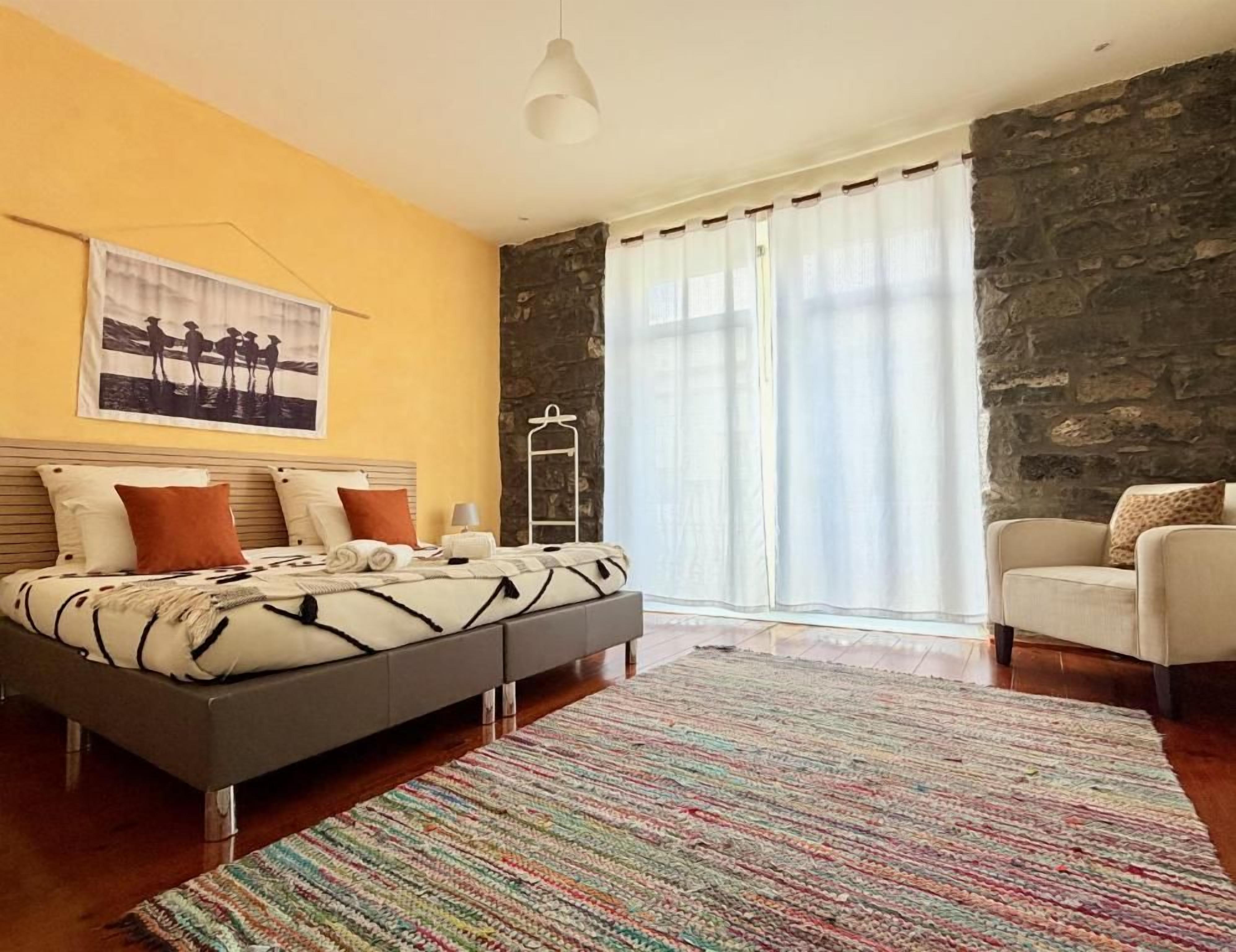 Un dormitorio con cama, una silla, una pared de piedra y una ventana con cortinas.