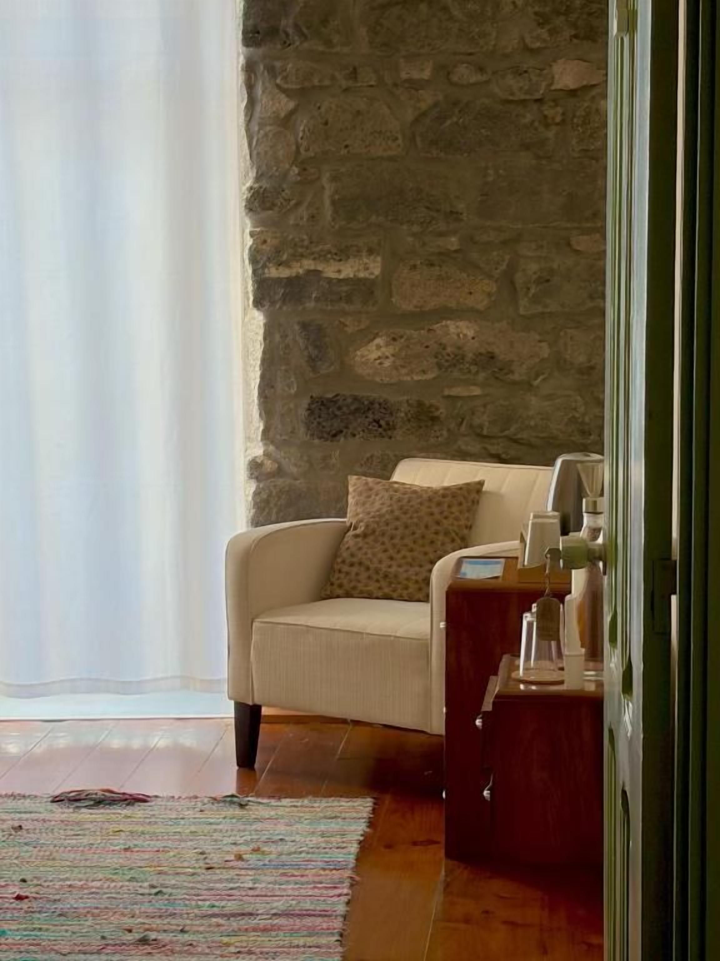Ein Zimmer mit einer Steinsäule, einem weißen Sessel mit gemustertem Kissen, einem Holztischchen mit Lampe und einem bunten Streifen Teppich.