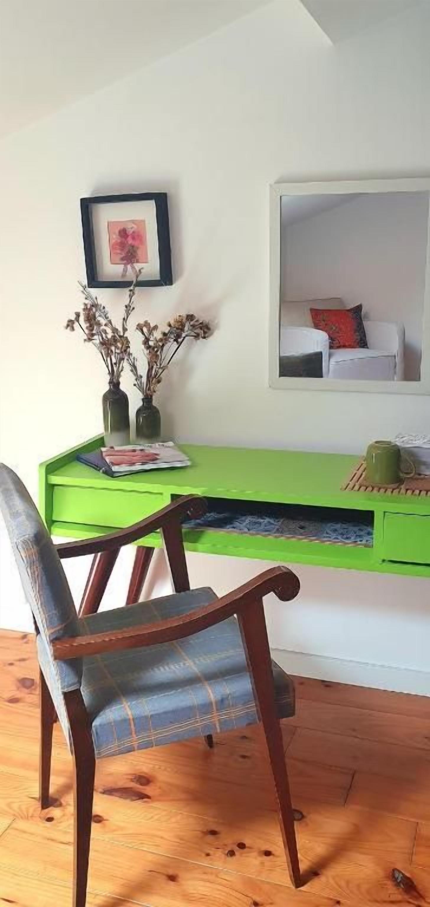 Una habitación con un escritorio verde, una silla, un cuadro enmarcado y un espejo que refleja una cama.