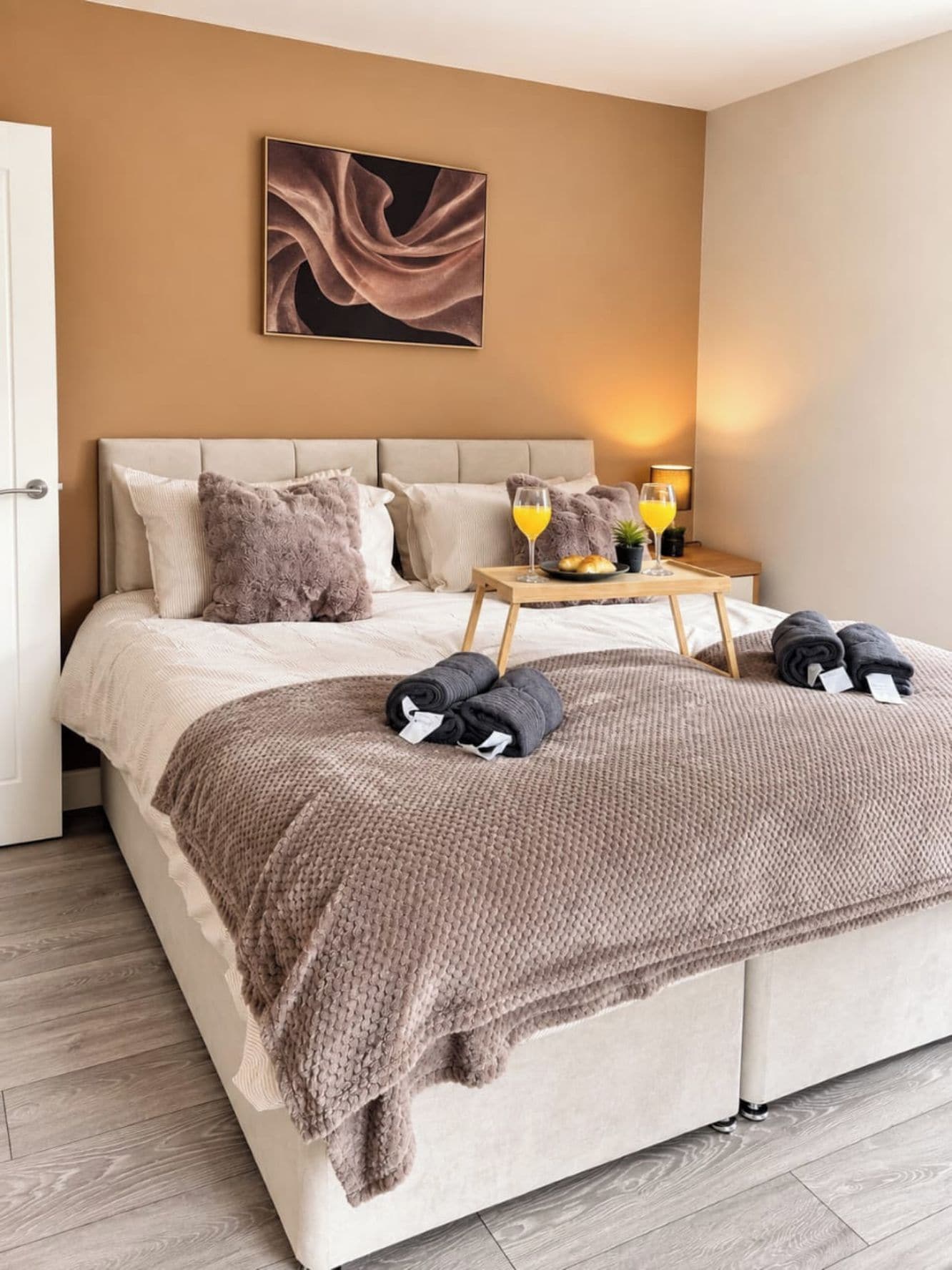 Un lit bien fait, avec une couverture marron, des oreillers et des serviettes enroulées. Sur une table de chevet, on trouve des verres de jus d’orange et une assiette de croissants. Un tableau est accroché au mur, au-dessus du lit.