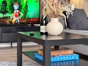 Un soggiorno moderno con una TV a schermo piatto che mostra un programma, una bottiglia di champagne, un vaso con piante secche, un telecomando e una scatola del gioco Scrabble.