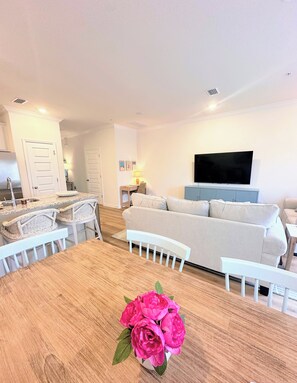 Una sala de estar moderna con una mesa de comedor de madera, sillas blancas, un sofá color beige y un televisor de pantalla plana montado en la pared.