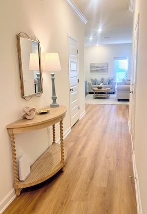 A hallway with a wooden console table, a mirror, and a lamp. There is a door leading to a living room with a sofa and a painting on the wall. - Brand new 3 bed/2.5 bath townhome central to the beach, Wharf, and restaurants! (Orange Beach)