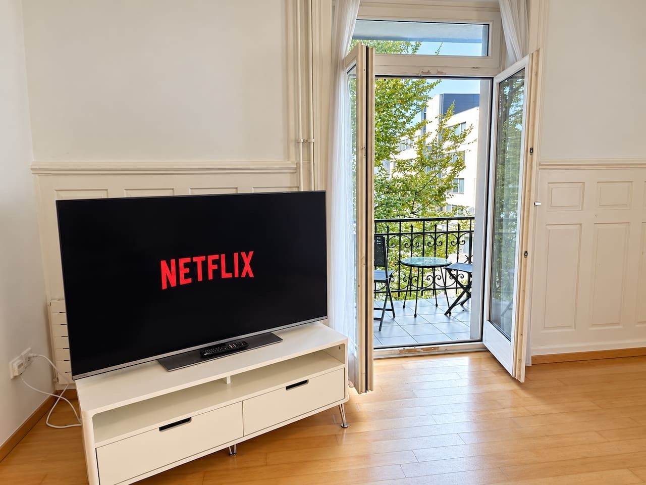 Une pièce avec un téléviseur affichant le logo de Netflix, une porte-fenêtre coulissante donnant sur un balcon extérieur équipé d’une table et de chaises, et un mur blanc.