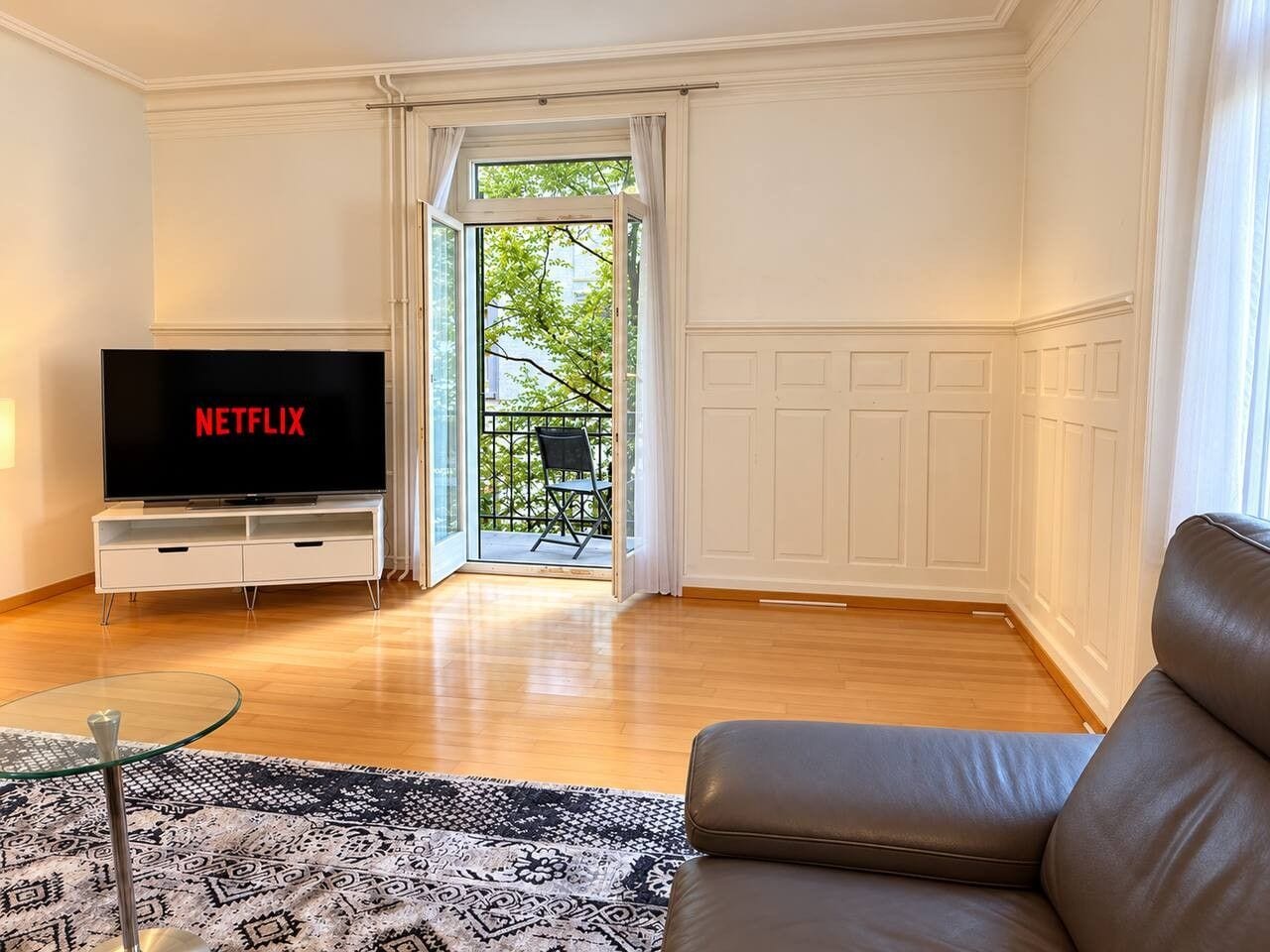 A living room with a flat-screen TV, a glass coffee table, a patterned rug, a leather sofa, and a sliding glass door leading to an outdoor area.