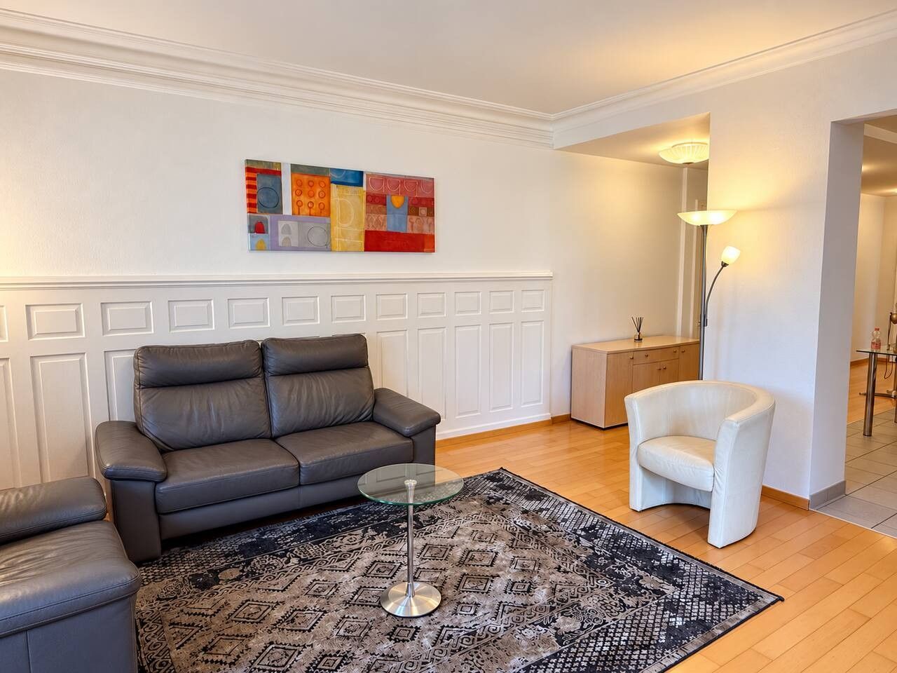 Un salon avec un canapé gris, une table basse en verre, un tapis à motifs, un fauteuil blanc et un tableau accroché au mur.