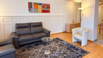 Un salon avec un canapé gris, une table basse en verre, un tapis à motifs, un fauteuil blanc et un tableau accroché au mur.