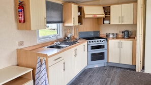 A compact kitchen with a gas stove, oven, and a sink with a dish rack. There is a fire extinguisher mounted on the wall, and a window with blinds. - Ingoldmells 2-bed Cabin, Sleeps 6 With Parking (Skegness)