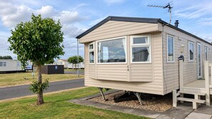 A single-story mobile home with a flat roof, a small porch, and a window with a curtain. - Ingoldmells 2-bed Cabin, Sleeps 6 With Parking (Skegness)