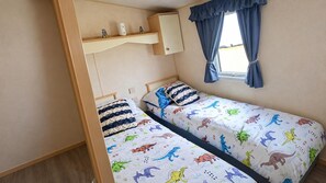 A single bed with a dinosaur-themed bedspread, a small shelf with toys, and a window with curtains. - Ingoldmells 2-bed Cabin, Sleeps 6 With Parking (Skegness)