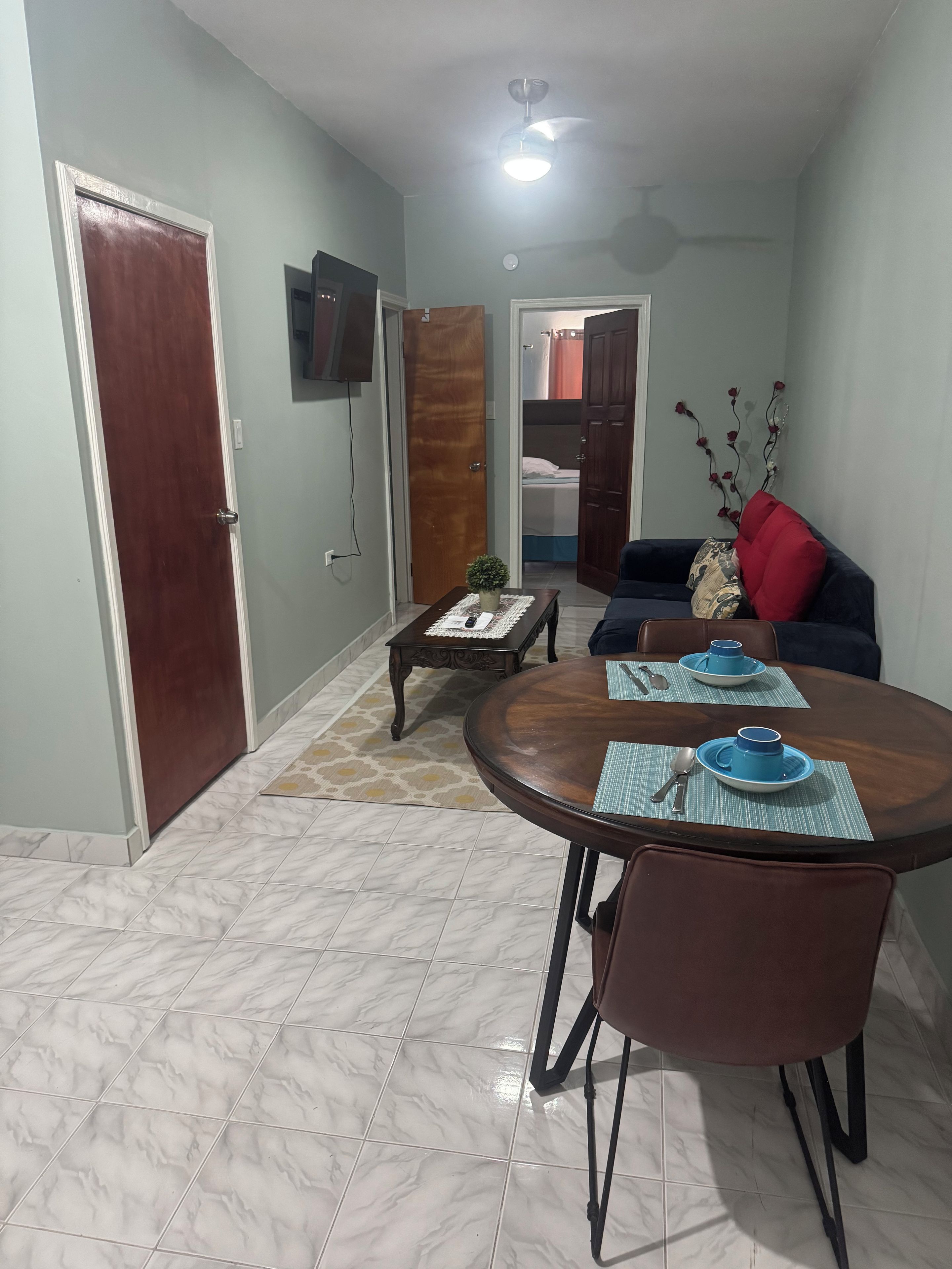 A dining area with a round table, chairs, and blue tableware. There is a sofa with a red pillow and a decorative branch with flowers. A television is mounted on the wall.