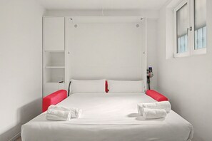 A single bed with white bedding, red pillows, and two white towels. There is a small shelf unit to the left of the bed, and a window with a view of the outside. - Apartment 'Smart Hub M3 Metro 10 Min.Duomo' with shared garden, Wi-Fi and air conditioning (Milano)
