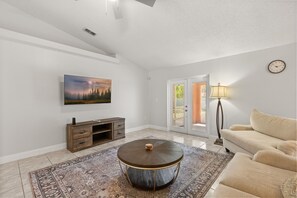 A living room with a round coffee table, a sofa, a TV mounted on the wall, and a door leading to an outdoor area. - Charming Kissimmee Retreat Near Orlando (Kissimmee)
