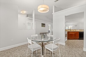 A dining area with a glass table and white chairs, a kitchen with wooden cabinets, and a ceiling light fixture. - Charming Kissimmee Retreat Near Orlando (Kissimmee)
