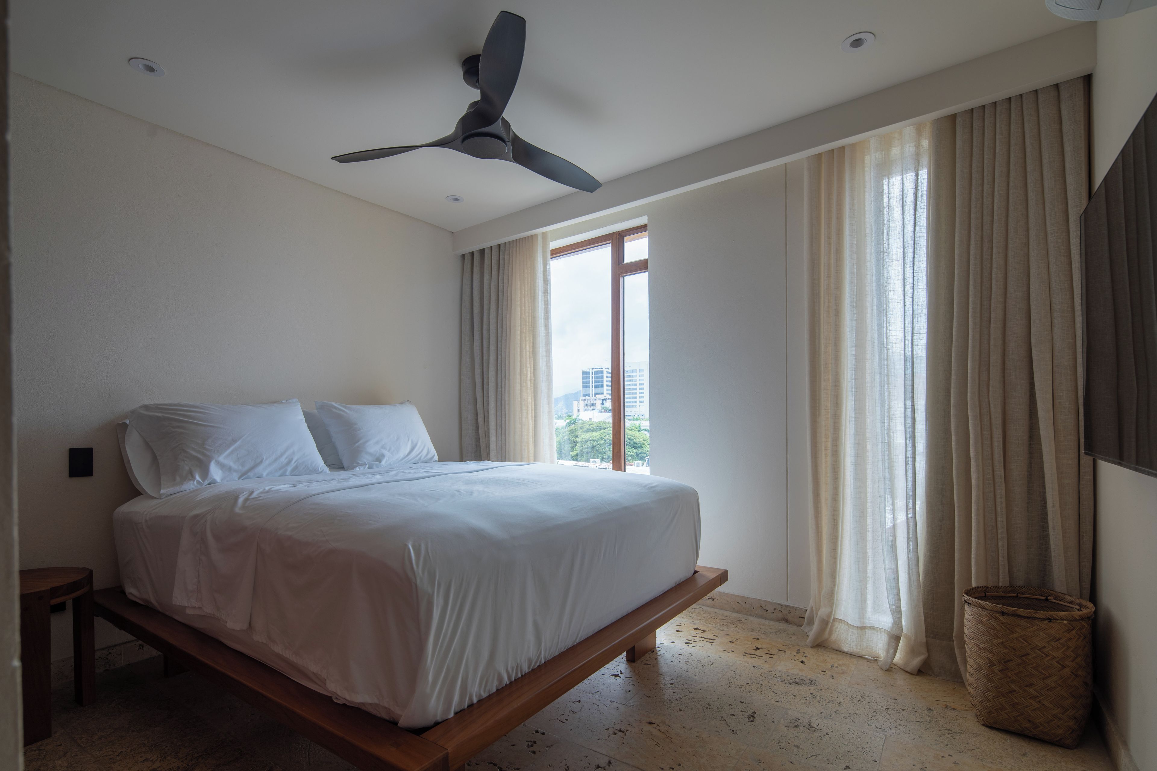 Ein Schlafzimmer mit einem Bett, einem Deckenventilator, einem Fenster mit Vorhängen und Blick auf Gebäude.