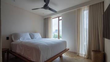 Ein Schlafzimmer mit einem Bett, einem Deckenventilator, einem Fenster mit Vorhängen und Blick auf Gebäude.