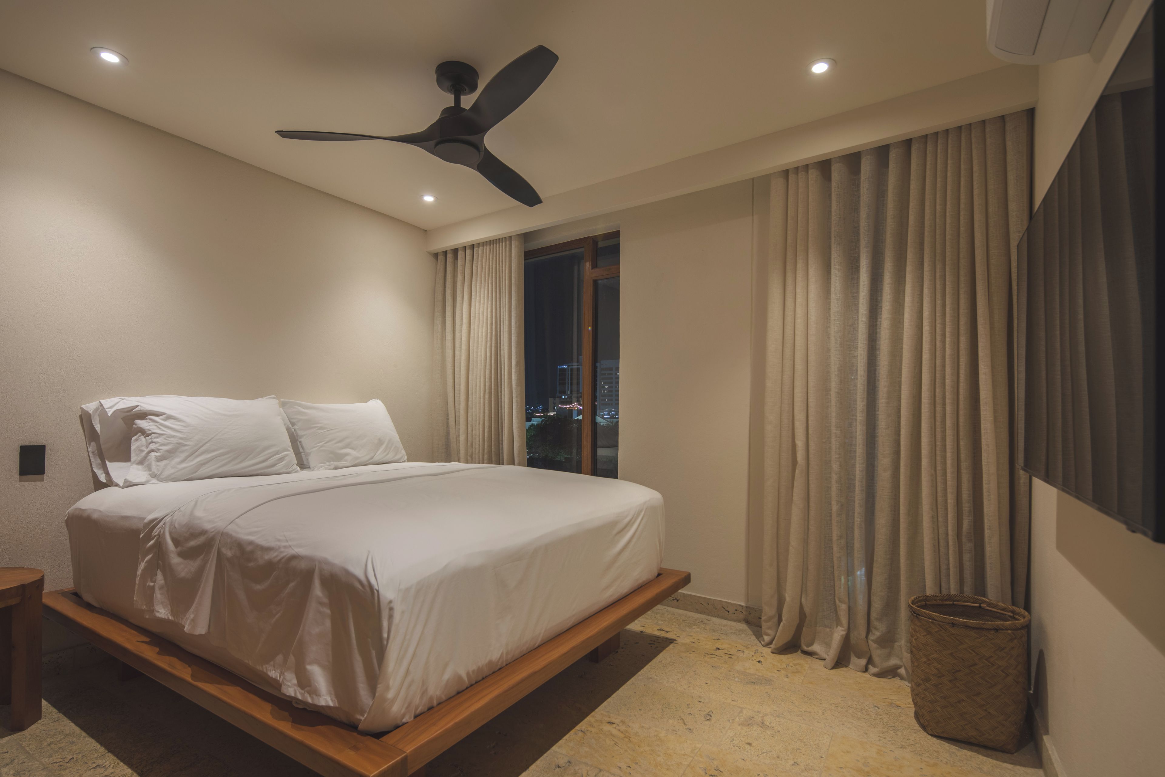 Ein modernes Schlafzimmer mit Bett, Deckenventilator und Blick auf die beleuchtete Stadt bei Nacht.