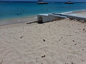 A sandy beach with a concrete structure, likely a lifeguard stand, and calm turquoise waters with boats in the distance. - Budget bungalow across the road from the calm west coast sandy beach (Paynes Bay Beach)