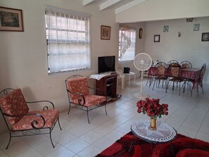 A living room with a TV, two chairs, a dining table with chairs, a fan, and a vase of flowers. - Budget bungalow across the road from the calm west coast sandy beach (Paynes Bay Beach)