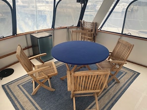 A round table with four wooden chairs, a blue tablecloth, and a blue and beige striped rug on a deck with a view of the water.