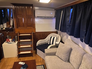 A compact living space with a sofa, a wicker chair, a small table, a refrigerator, a wooden cabinet, a ladder, and an air conditioning unit.