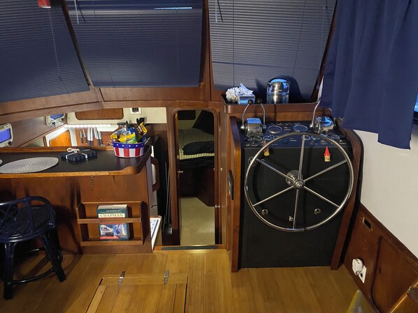 A compact galley with a steering wheel, a small sink, and a shelf with books and snacks.