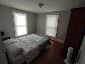 Un dormitorio con cama, dos ventanas con persianas, suelo de madera y un radiador.