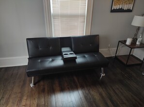 Ein schwarzes Ledersofa mit einer zentralen Konsole, vor einem Fenster mit Jalousien platziert, auf Holzdielen, mit einem kleinen Beistelltisch und einer Lampe zur Rechten.