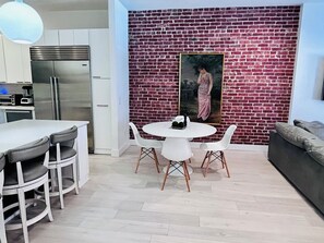 Une cuisine moderne avec un réfrigérateur en acier inoxydable, des armoires blanches et un mur en briques orné d’un tableau encadré.