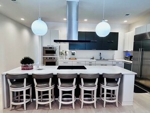 Une cuisine moderne dotée d’un îlot central, de tabourets de bar, d’appareils électroménagers en acier inoxydable et de luminaires suspendus.