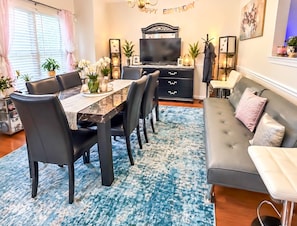 A dining area with a table, chairs, and a sofa. There is a television on a stand, a bookshelf, and a wall-mounted shelf with decorative items.