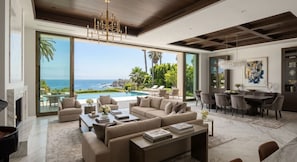 A spacious living room with a large window overlooking a pool and ocean, furnished with sofas and a dining area. - Exceptional Homes for  Exceptional Lives (Phoenix)