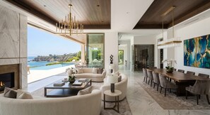 A modern living room with a fireplace, a dining area with a wooden table and chairs, and a large window overlooking a pool and ocean view. - Exceptional Homes for  Exceptional Lives (Phoenix)