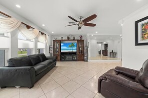 A spacious living room with a large flat-screen TV, a ceiling fan, and a view into the adjacent dining area. - The Pool House | Spa, Game Room & Home Gym (Cape Coral)