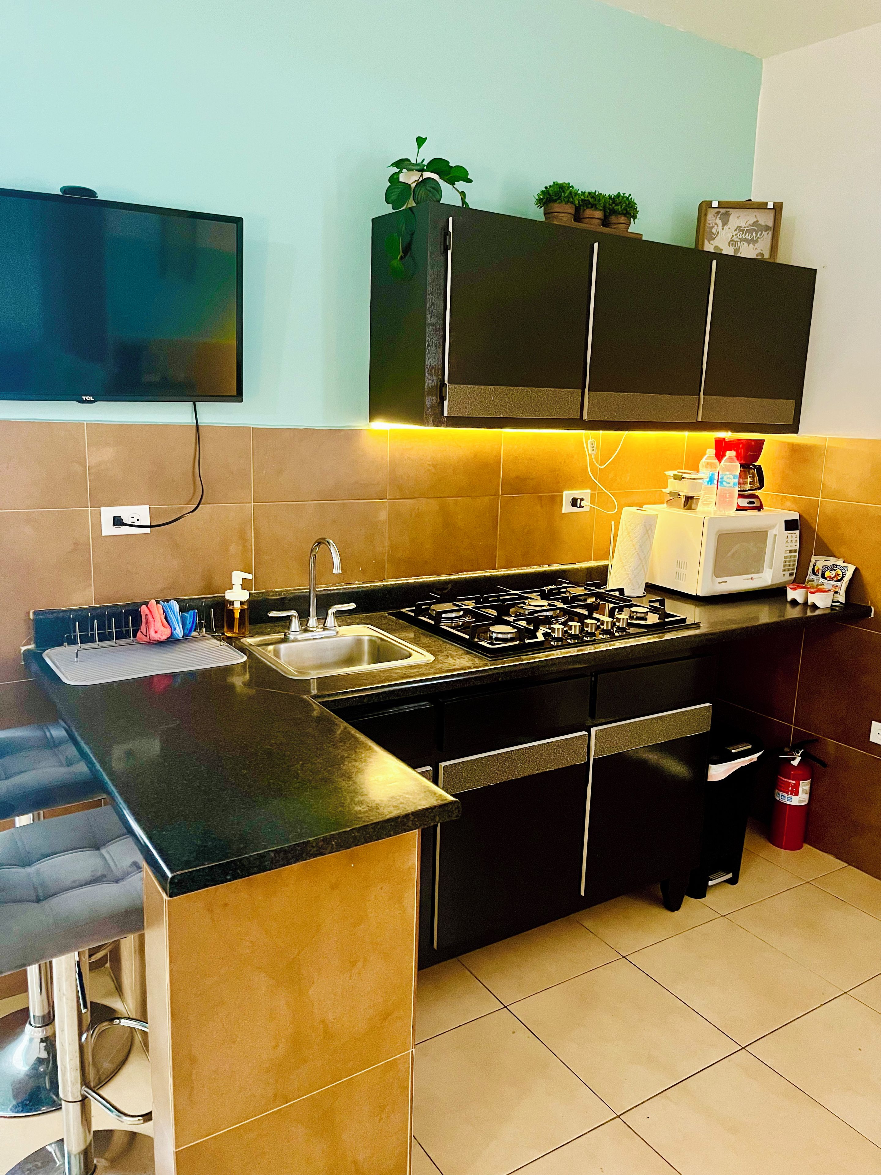 A modern kitchen with a gas stove, sink, and a microwave. There is a bar counter with stools and a television mounted on the wall.