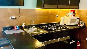 A modern kitchen with a gas stove, sink, and a microwave. There is a bar counter with stools and a television mounted on the wall.