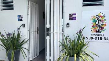 Entrada de hotel con una reja blanca, plantas en macetas y un letrero con un número de teléfono.