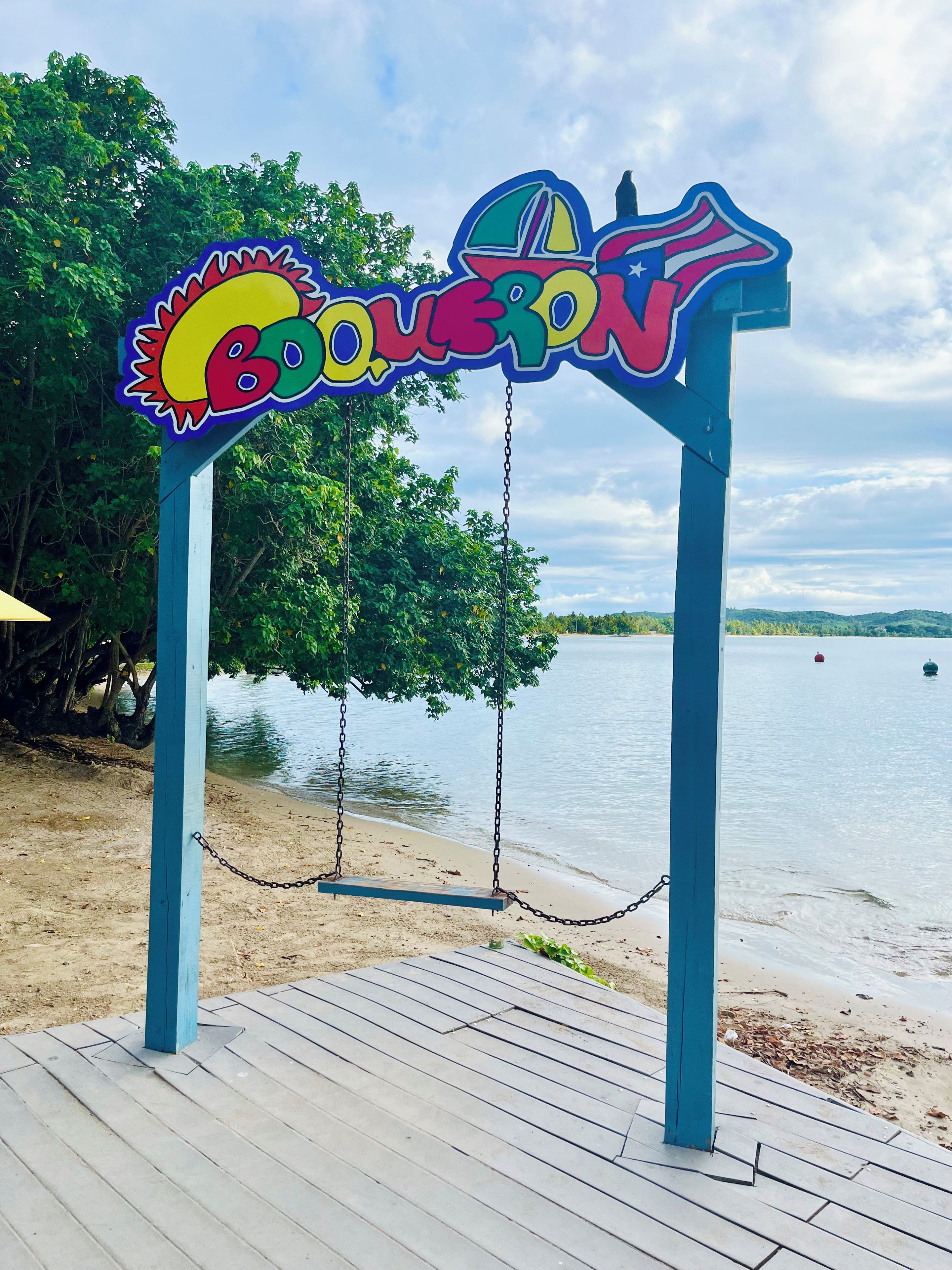 Un espace de jeux avec des balançoires situé au bord de la plage, agrémenté d’une enseigne colorée, d’une terrasse en bois et offrant une vue sur l’eau.