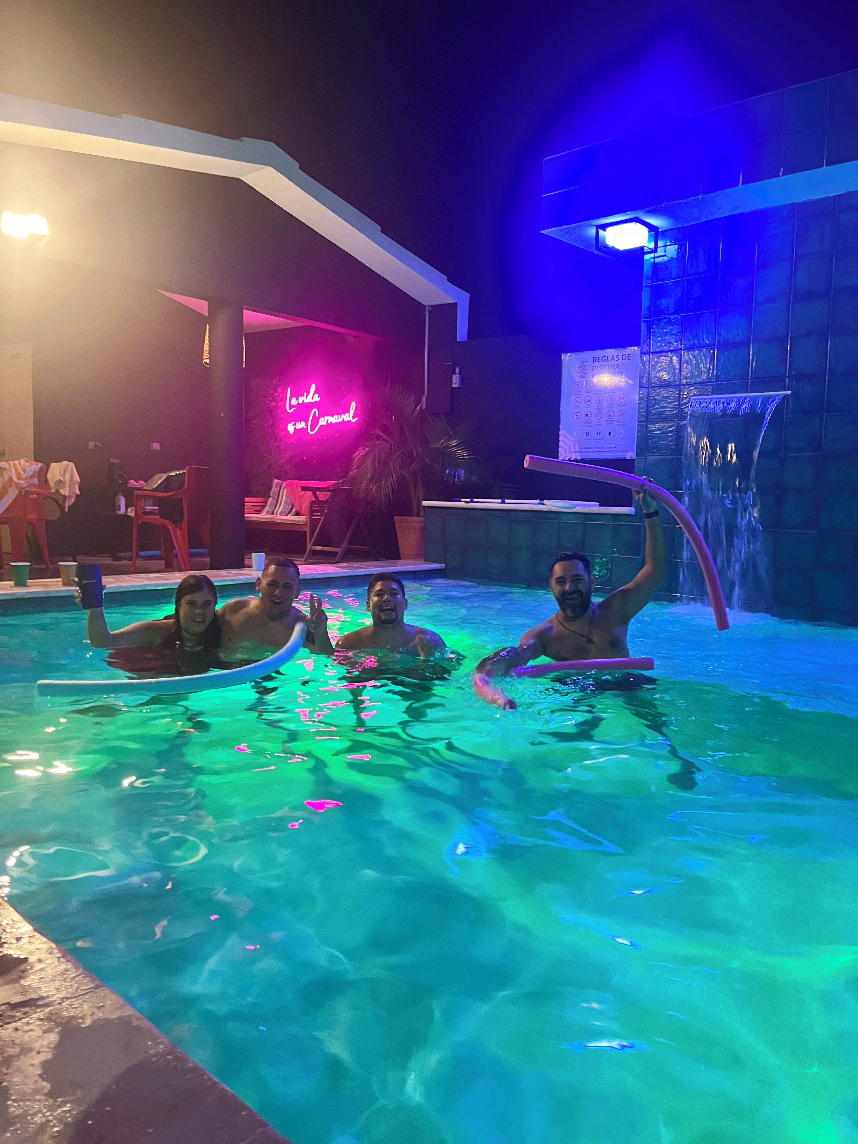 A swimming pool area with a neon sign, a waterfall feature, and a poolside seating area.