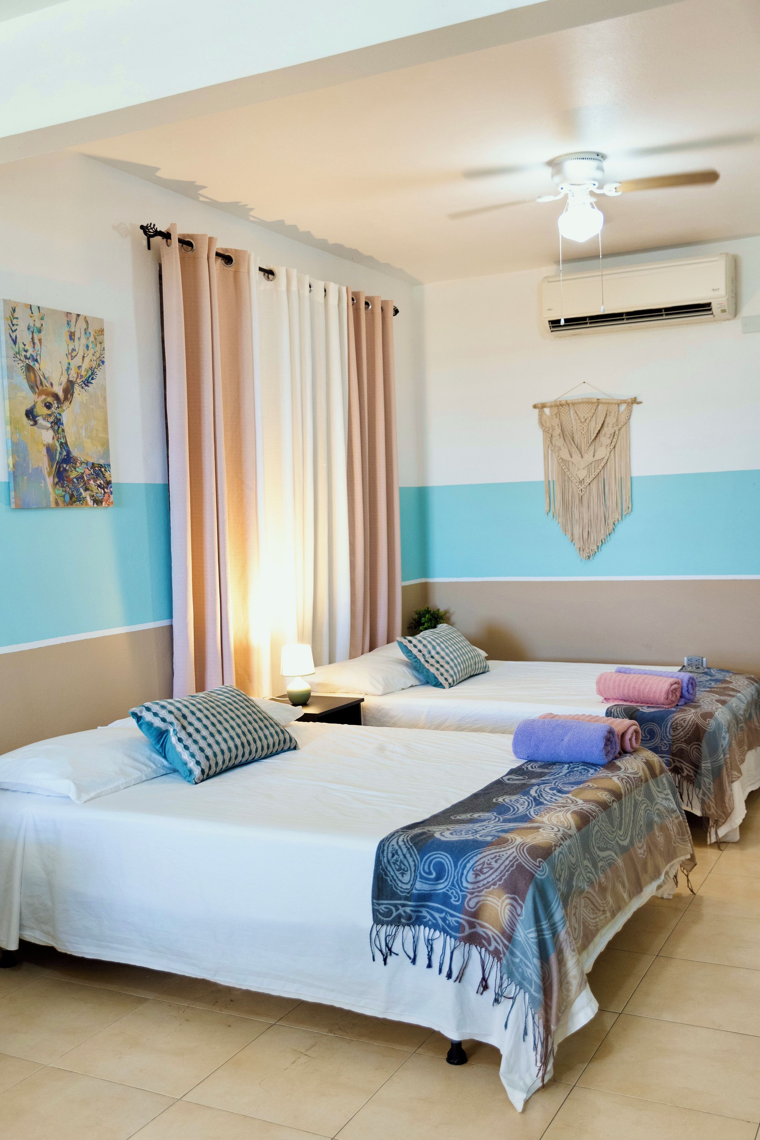 A hotel room with two beds, a ceiling fan, an air conditioning unit, and a wall-mounted macrame decoration.