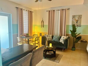 A living room with a sofa, a dining table with chairs, a lamp, and a painting on the wall. - Mountain Retreat with Boquerón Bay Views & Heated Saltwater Pool - 3B (Cabo Rojo)