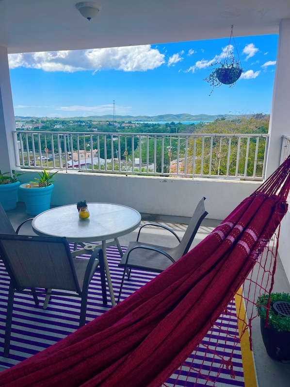Un balcon avec vue sur le paysage urbain, une table ronde avec des chaises, un hamac et des plantes en pot.