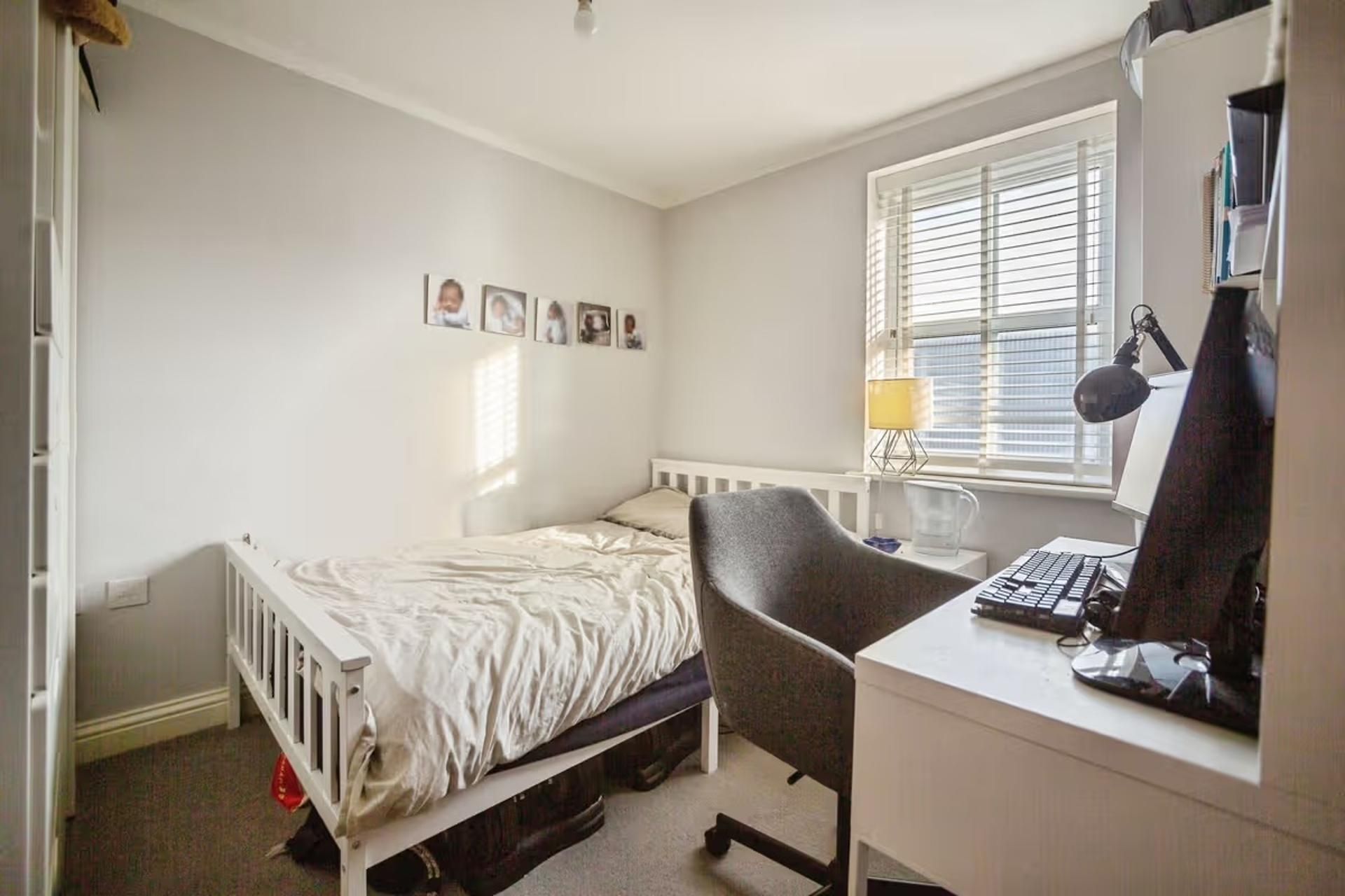 A bedroom with a single bed, a desk with a computer, a chair, and a window with blinds.