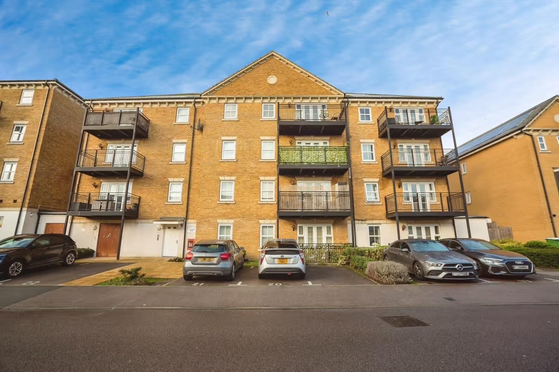 A multi-story residential building with balconies, a parking area with cars, and a clear sky.