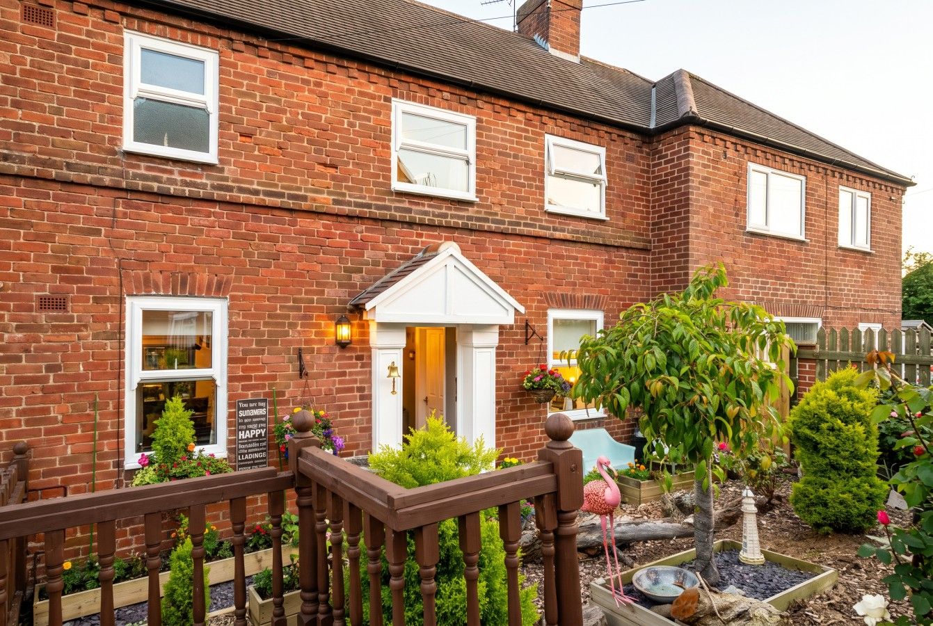 A brick house with a front porch, a small garden with a flamingo, and a wooden fence.