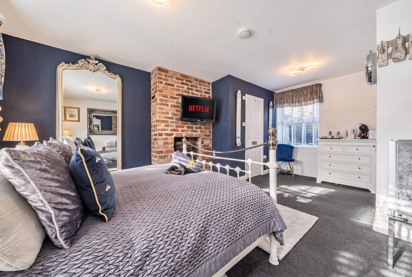 A bedroom with a bed, a TV mounted on a brick wall, a dresser, and a window with curtains.